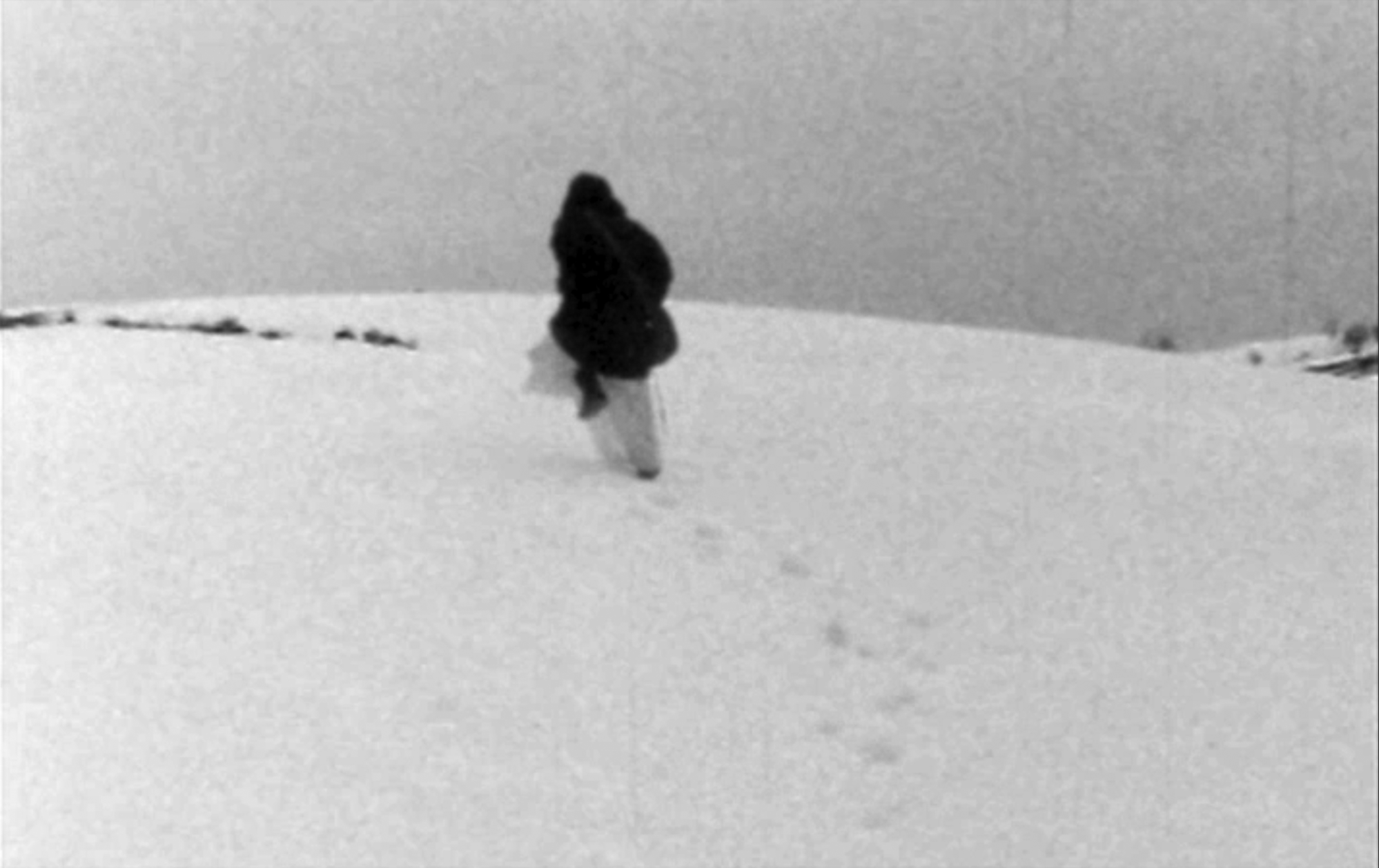 black and white image, a person trudges up a snow covered hill, their back is towards us